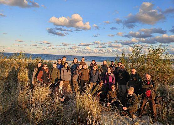 Konferencja INVENT 2025 Ludzie w wysokiej trawie przy morzu, otoczeni naturalnym krajobrazem i błękitem wody.